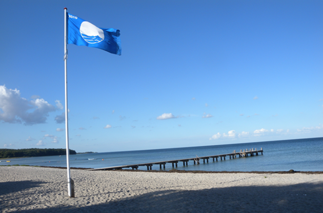På et billede ses en strand med en bådebro og en flagstang med et blåt flag.
