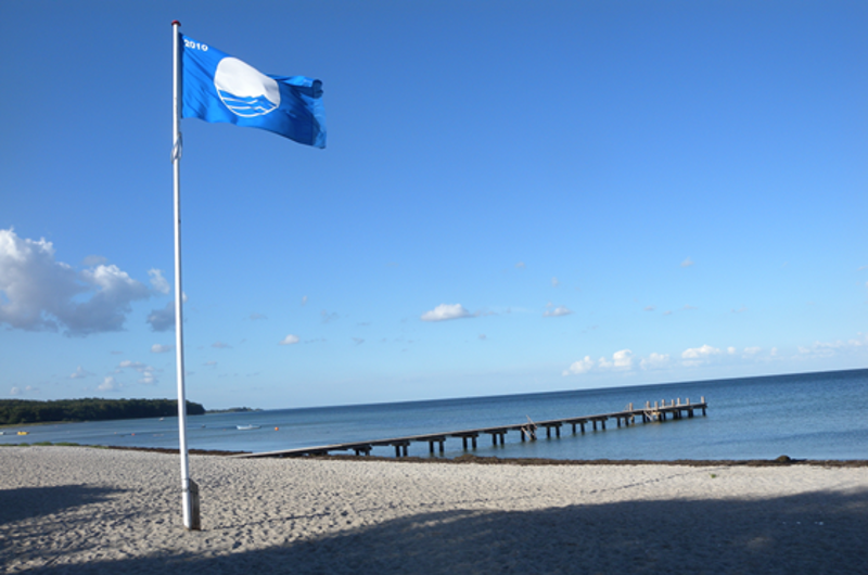 På et billede ses en strand med en bådebro og en flagstang med et blåt flag.