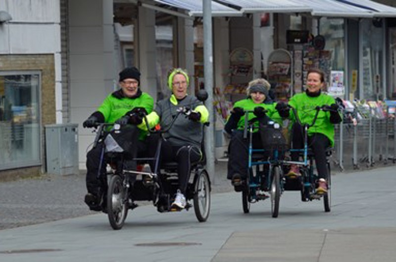 På billedet ses to cykler, der hver cykles af to personer som sidder ved siden af hinanden. De er på en gågade og der ses personer som går i baggrunden.